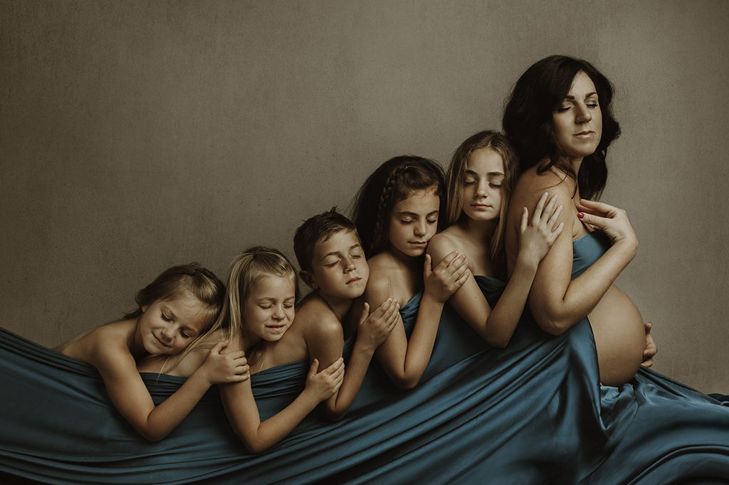 Foto artistica a colori di famiglia con le sole donne, mamma e le figlie dalla più grande alla più piccola, con un fratello, tutti abbracciati. Tutte coperte da un velo in raso di colore verde. Fotografia Fine Art di famiglia a colori Trieste