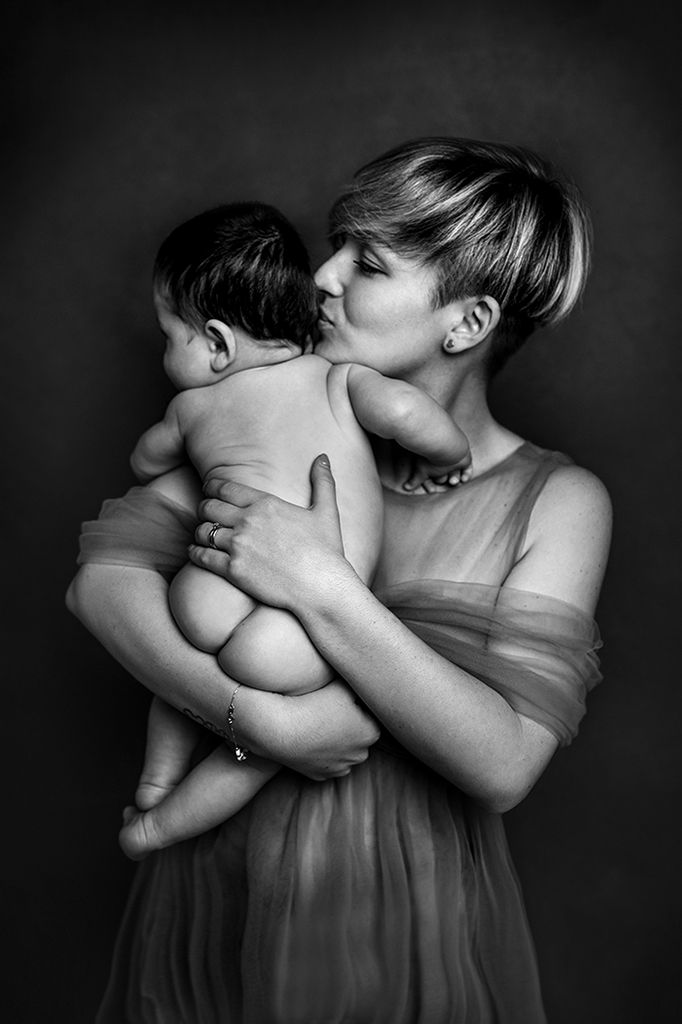 Foto ritrattistica emotional in bianco e nero con mamma che tiene in braccio il neonato a cui dà un bacio sulla guancia. Fotografia emotional in bianco e nero Trieste