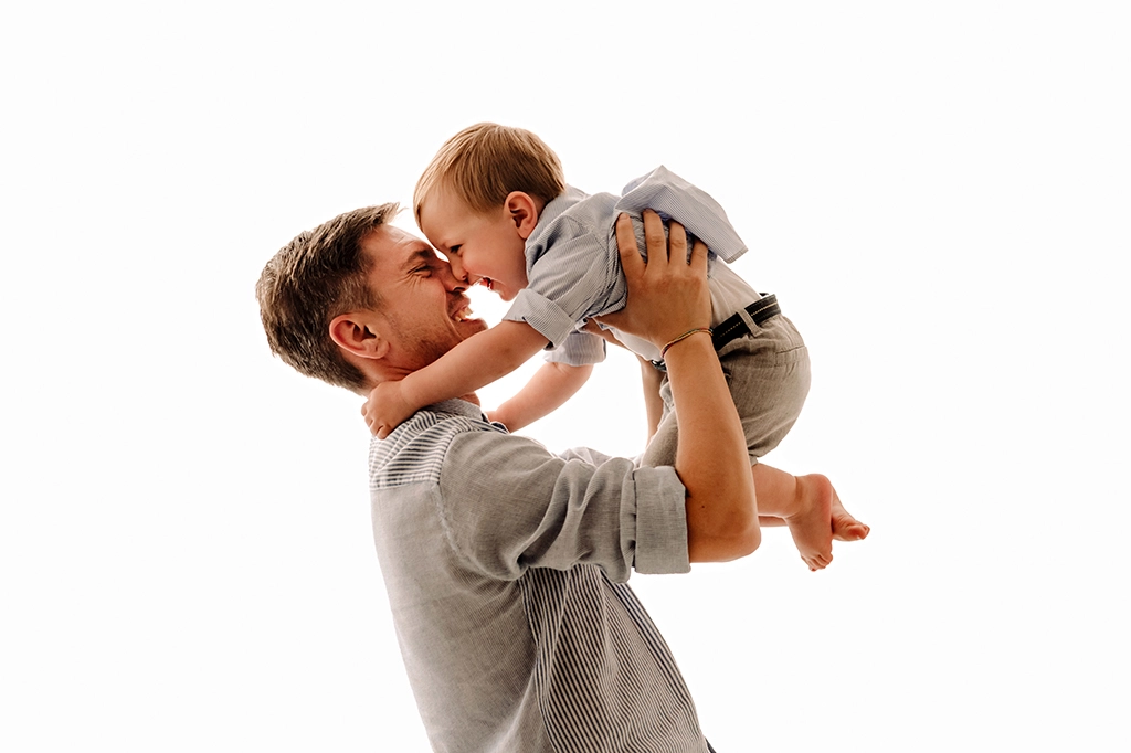 Foto a colori in studio di famiglia, mentre il papà tiene in braccio il bambino e si guardano ridendo, naso contro naso. Fotografia a colori di famiglia papà e bambino Trieste