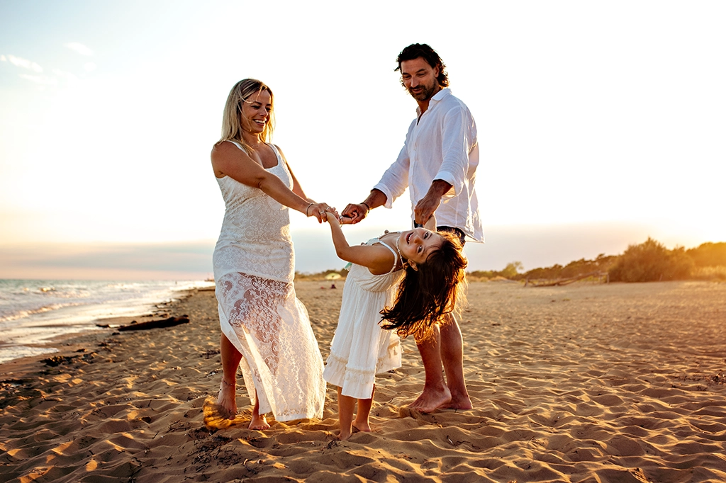 Foto fuori studio a colori di una famiglia sulla spiaggia che si tiene per mano al tramontare del sole, tutti e tre vestiti di bianco. Fotografia fuori studio a colori Italia