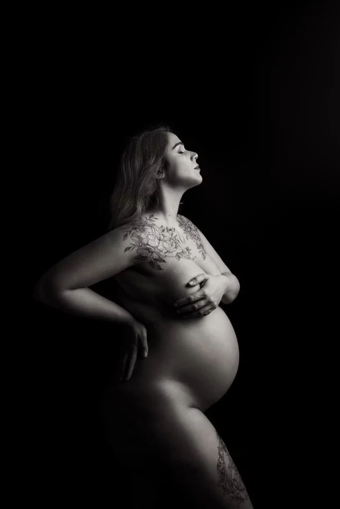 Foto in bianco e nero in studio di maternità, di una donna incinta tatuata nuda, mentre si regge il seno. Fotografia a colori in studio di maternità Trieste